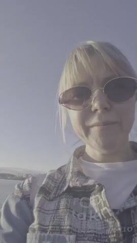 The video shows a calm lake under a bright sun. A woman with blonde hair and sunglasses is visible in the initial frame, partially obscuring the view. The camera then shifts to focus on the lake and the sun, which is shining brightly and reflecting off the water's surface. There's a hint of land in the distance, appearing as a dark line on the horizon. The sky is clear and pale blue. The overall tone of the video is peaceful and serene.