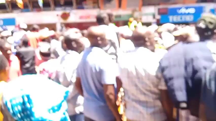 Alright, here's a detailed description of the video:

The video captures a large crowd of people gathered outdoors in what appears to be a city or town setting. The majority of the individuals are facing the same direction, presumably towards a focal point of interest. The crowd is densely packed, and people are wearing a variety of clothing, including shirts, jackets, and hats. There are signs visible, and banners in the background, suggesting a possible political rally or public event. Buildings line the street behind the crowd, featuring shops with visible signage and outdoor seating areas. The weather appears to be sunny. The camera moves through the crowd, providing different perspectives of the gathering. The overall impression is of a vibrant, bustling atmosphere with a strong sense of collective energy.