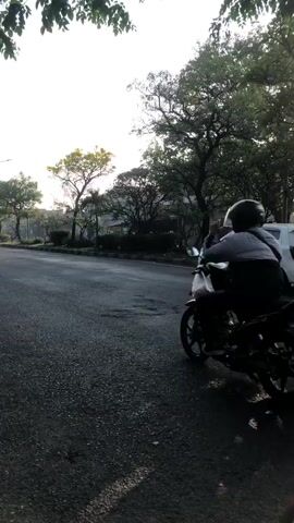 A sunny day with some light and shadows suggests it is either early morning or late afternoon. The sky is bright white and slightly hazy, indicating a cloudless day. Green leaves and branches are visible at the top of the frame, casting dappled shadows on the asphalt road. The road itself appears to be in decent condition with some small patches of rougher texture.

In the foreground, the camera is positioned low to the ground, giving a perspective from a vehicle. Several motorcycles and a light-colored car are present on the road, moving in the same direction. The vehicles are spread out, with some closer to the camera and others further away.

On the left side of the road, two figures are riding on motorcycles. The first motorcycle, in the middle ground, has two people on it. The person in the front is wearing a white helmet, and the person in the back is wearing a dark helmet. The second motorcycle, closer to the foreground, has one person on it wearing a dark helmet.

Further down the road, a white car is visible. Behind it, two more motorcycles are seen. The motorcycle on the right has two people on it, both wearing helmets. The motorcycle on the left has one person wearing a helmet and a backpack.

In the background, a large blue and silver dump truck is moving. It appears to be traveling in the same direction as the other vehicles. More trees and some buildings are visible beyond the road. The overall impression is a typical urban street scene with moderate traffic.