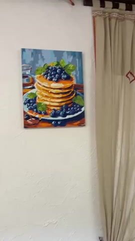 The video opens with a close-up of a painting of pancakes with blueberries. The camera then pans to the left, revealing another painting of flowers. The video continues to pan, showing a colorful crocheted blanket hanging on a white wall. Two people, a woman wearing a blue baseball cap and a man wearing a gray baseball cap, are seated at a wooden table. The woman appears to be looking at her phone, and the man is looking down at the table. The camera then pans to the right, showing a window and a shelf with framed pictures and decorative items. The video finishes with a shot of a wooden table with placemats and napkins, and a window in the background. The environment appears to be a dining area or a cafe.