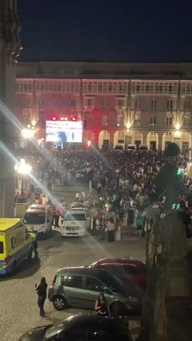 The video shows a crowded public square at night. A large screen is set up at one end, displaying an event, and a crowd of people are gathered in front of it, facing the screen. Several vehicles, including an ambulance and police cars, are present in the foreground, near the crowd. Buildings with balconies line the square. People are walking around, some are holding drinks, and others appear to be watching the screen. The lighting suggests it is evening or night.