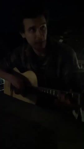 A man is playing an acoustic guitar in a dimly lit environment. He is wearing a dark shirt and is seated with the guitar resting on a surface in front of him. The lighting is poor, making details hard to see, but the guitar is light-colored with a dark pickguard. The background is mostly dark, with vague suggestions of structure or foliage.