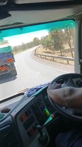 A driver navigates a truck on a winding road. The perspective is from inside the cab, showcasing the driver's hands on the steering wheel and the dashboard. Ahead, another truck with a covered bed is visible, moving in the same direction. The road curves to the left and is bordered by a metal guardrail, with dry, grassy terrain and scattered trees beyond. The sky is bright, suggesting a sunny day. The driver seems to be focused on the road and maintaining a safe distance from the truck ahead.