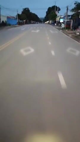 The video shows a first-person perspective of riding a vehicle down a paved road during twilight. The road has multiple lanes marked with white lines, including a double yellow line on the left side. Road markings indicate lanes for going straight and turning left. A pedestrian crossing is visible. On either side of the road, there are trees, buildings, and some signs. The sky is dark blue, suggesting it is either early morning or late evening. The video is shaky, indicating movement. There are no people clearly visible in the frame.