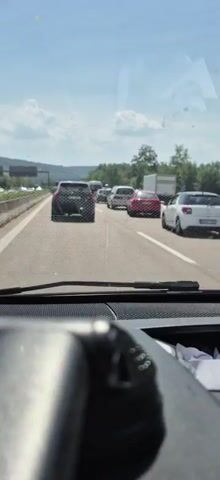 A video recorded from inside a vehicle shows traffic congestion on a highway. The view is through the windshield, with the dashboard in the foreground. Several cars are visible on the multi-lane road, moving slowly or stopped. The sky is bright and appears to be daytime, with some clouds. Trees and hills are visible in the background. The road surface is asphalt. The cars are of various colors, including black, white, and red. The overall impression is that of a traffic jam.