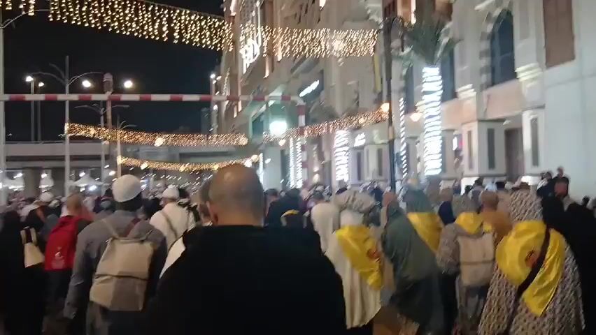 A dense crowd of people moves down a street at night, under bright rows of festive lights hung overhead. The lights are arranged in parallel lines across the street, and some include crescent moon symbols. The buildings lining the street are brightly lit and have a distinctive architectural style. A metal structure with red and white striped poles spans the street. Many people are wearing white caps. Some individuals are wearing yellow or green sashes. The scene has a festive and crowded atmosphere.