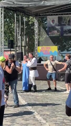 A group of people are gathered outdoors in front of a stage. A man in traditional white clothing with a sash is singing into a microphone. He is standing next to a woman in a long blue dress. To their left, a man in a black vest and jeans is holding a microphone and dancing with a woman in a floral dress. Further left, a man in a black shirt and hat is also holding a microphone. In the background, there is a banner with text in yellow and blue. To the right of the stage, a man in a light-colored polo shirt and shorts is walking and holding a bottle of water. Another couple is dancing to the right of him. The ground is paved with cobblestones. Trees and a body of water are visible in the background behind the stage. The sky is clear and sunny.