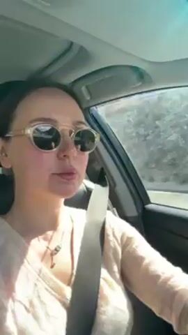 A woman with light skin and brown hair, wearing round sunglasses and a beige sweater, drives a car along a highway through a mountainous region. She is wearing a seatbelt and has a necklace with a small pendant. The car's dashboard is visible, with a navigation system and other controls.  The landscape outside the car window features gray, rocky mountains under a clear blue sky. Another car can be seen on the highway ahead. The overall tone is casual, suggesting a road trip or everyday commute through a scenic area.