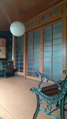 The video shows a porch with wooden flooring, sliding glass doors, and a hanging paper lantern. There are two benches and a small table near a stone wall, decorated with a painting of cranes.  A pond with various plants and rocks is located next to the porch. The overall aesthetic is a mix of Japanese and garden decor. The camera pans from right to left, showing more of the garden and pond. The sound of the wind can be heard in the background.