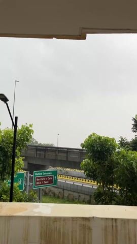 From a low-angle vantage point, a view of a freeway under cloudy skies unfolds. The camera is positioned behind a short, pale-colored wall that cuts across the lower portion of the frame. Lush green trees flank the sides, framing the road scene.

A prominent green sign provides directions to Mutiara Damansara, pointing towards the Curve and IKEA/Tesco. Overhead, a concrete overpass stretches across the freeway, while numerous vehicles, including cars and trucks, are visible traveling along the road. Streetlights are interspersed along the side of the freeway. The overall impression is that of a cloudy day in a busy suburban or urban area.