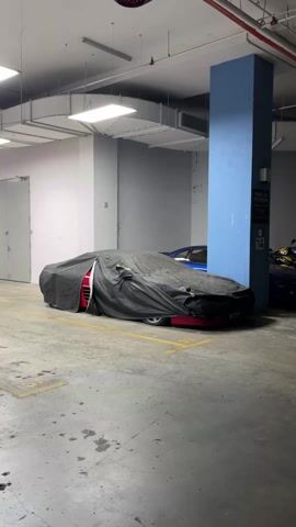 In an indoor parking garage with fluorescent lighting and overhead pipes, a red car is parked under a black car cover. A blue car is parked beside it. Adjacent to the blue car are two motorcycles. There is a blue support column on the right of the frame. A closed metal door is on the left. The floor is concrete with yellow painted parking lines.