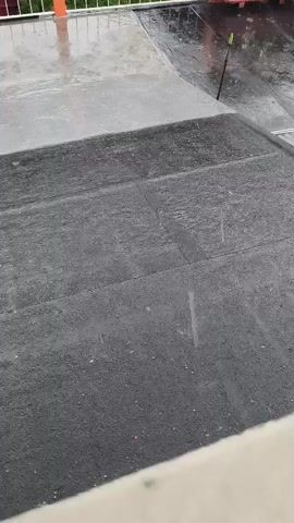 The video captures a flat rooftop during a rain shower in Quito, Ecuador. The foreground of the roof is covered with a dark, textured material, possibly a gravel-surfaced or granular-modified bitumen membrane, showing distinct seam lines. The background section of the roof appears smoother and lighter in color, possibly concrete or a different type of membrane, with water visibly pooling on its surface.

Heavy rain is falling consistently, creating splashes and causing water to flow across the roof's surface. Water has accumulated in various depressions, reflecting the overcast sky. A white railing is visible along the perimeter of the roof in the background, suggesting a boundary or a balcony. Partially visible on the left side of the frame is an orange vertical structure, possibly a pipe or part of an awning support. A dark, thin stick or tool is seen standing upright in a pool of water on the lighter-colored roof section towards the right. No people or visible text are present in the video. The overall scene depicts a functional rooftop enduring a significant downpour.