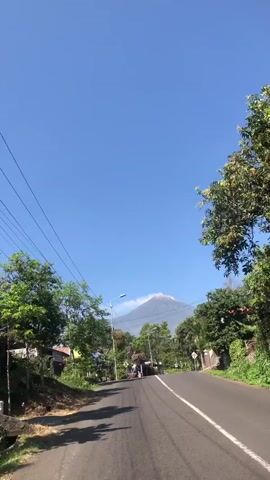 The video shows a view from a moving vehicle on a paved road, likely in a rural area given the presence of trees and modest buildings. The sky is clear and blue. In the distance, a large mountain, possibly a volcano, is visible, with its peak partially obscured by clouds. The road has a white line marking the center and edges. Along the sides of the road are green trees, some utility poles with wires, and a few houses or buildings. There are a couple of motorcycles and a white car visible on the road, suggesting some traffic. The overall impression is of a sunny day in a scenic location with a prominent natural landmark.