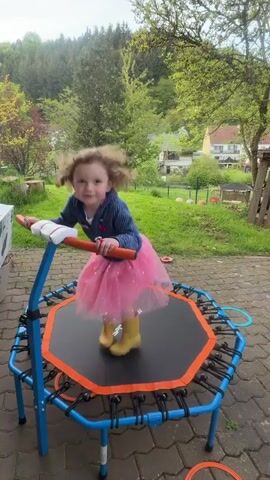 A young girl with curly hair, dressed in a blue long-sleeved top, a pink tutu, and yellow wellington boots, is joyfully jumping on a small, octagonal trampoline. The trampoline has a blue metal frame, orange trim, and a black mat with the word "SPORTDAY" printed on it. She holds onto an orange and white handlebar for stability.

The scene takes place outdoors on a paved surface, likely a patio or backyard in Trassem, Germany. In the background, there's a lush green lawn, various trees, and houses, suggesting a suburban or rural setting. The weather appears bright and sunny. The girl smiles and laughs while jumping, displaying clear happiness and excitement.