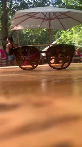 A pair of sunglasses rests on a wooden table in the foreground. The lenses reflect a scene of an outdoor seating area with people, tables, and a large white umbrella providing shade. Trees and foliage are visible in the background, suggesting a natural or park-like setting. The lighting indicates it is daytime.