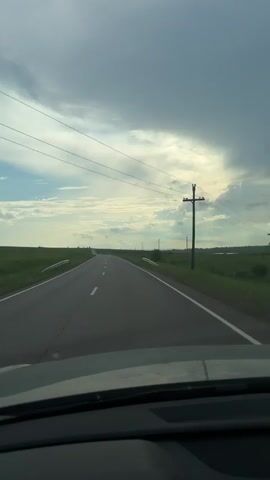 This video shows a point-of-view shot from inside a vehicle, looking forward through the windshield. The vehicle is driving on a paved road with white lines marking the lanes. The road stretches out into the distance, disappearing over a slight rise. On either side of the road are flat, green fields. Utility poles with wires are visible along the right side of the road. The sky is cloudy with patches of blue sky visible. The weather appears overcast, possibly with a storm approaching. The timestamp indicates the video is playing from 00:00 to 00:04.