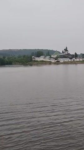 A drone shot captures a wide-angle view of a monastery on a distant shore across a body of water. The monastery complex features several white buildings with dark roofs and a prominent green-domed structure. To the left of the monastery, a line of trees borders the land. The water in the foreground is rippled, indicating a slight chop on the surface. The sky is overcast and grey.