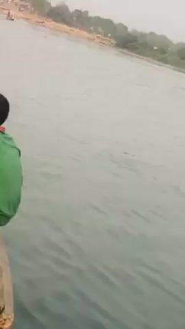 The video appears to have been taken near a river. The opening shot shows the back of a person with a silver head covering, a black top, and what seems to be a healing abrasion on their upper arm. Another person in a green shirt is partially visible. The view shifts to two men standing near the riverbank, one talking on a cell phone while holding a red and white object. The video then shows a wide shot of the river, focusing on the water's surface and the shoreline with trees and vegetation in the distance. The sky is overcast, and the water is a grayish-green color. Other boats are visible in the background in some shots.