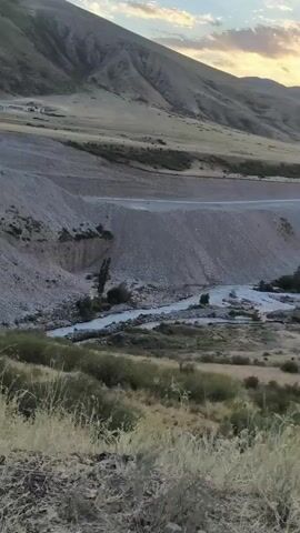 The video pans across a valley with a winding river. The landscape is dry and hilly, with sparse vegetation. There are some structures visible, possibly buildings or infrastructure, along the river and on the hillsides. The sky is partly cloudy with the colors of sunset visible, casting a warm glow over the scene. The overall impression is of a natural, somewhat remote environment during twilight.