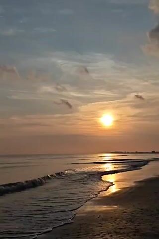 The video shows a sunset at the beach. The sun is low on the horizon, casting a golden glow over the water and sand. There are a few wispy clouds in the sky. The waves are gently lapping at the shore. No people are visible.