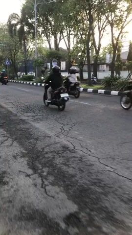 The video shows a street scene with several people riding motorcycles. The motorcycles are moving forward on a cracked asphalt road. The sun is setting and casts a warm glow on the scene. Trees line the side of the road, and there are buildings in the background. The people on the motorcycles are wearing helmets and casual clothing. One person is wearing a red shirt, and another is wearing a black jacket. The overall atmosphere is one of movement and activity.