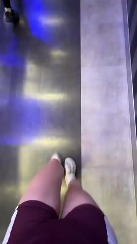 The video begins with a first-person perspective shot looking down at the legs of a person wearing burgundy shorts with white stripes and white sneakers, walking on a tiled floor with blue and yellow lighting. The view then shifts to a slightly higher angle as the person turns to face the camera. The person, a young woman with brown hair in two braids, is wearing a gray crop top and the same burgundy shorts and white sneakers. She is holding a bottle with a blue lid in her right hand. The background appears to be a gym or fitness center, with exercise equipment visible, including weight racks and machines. The woman smiles and nods at the camera as she continues to walk, turning to the right and looking around the gym. The video briefly returns to the original low-angle perspective of the person's legs walking before ending.