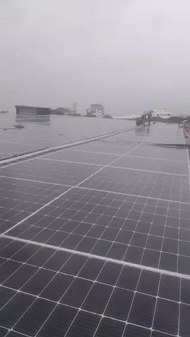 The video shows a close-up view of solar panels on a rooftop, with two people working on them in the distance. The sky is overcast and it appears to be raining. The environment is an urban setting with buildings visible in the background. One person is wearing a red shirt and the other appears to be wearing an orange shirt. They are both standing on the solar panels and seem to be performing some kind of maintenance or installation work. The solar panels are arranged in neat rows and cover a large area of the rooftop. The overall atmosphere of the video is one of hard work being done in challenging weather conditions.