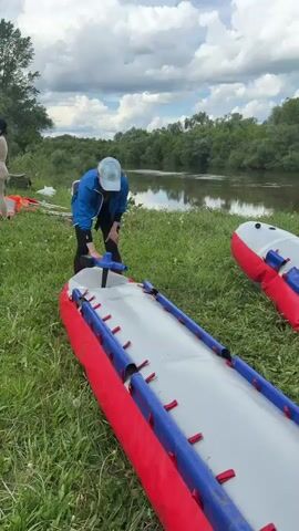 Two women are preparing inflatable kayaks next to a calm river. The first woman, wearing a blue jacket and cap, is inflating a kayak with a hand pump. The kayak is red and grey with blue accents. The second woman is kneeling over a similar kayak, adjusting something inside. The kayaks are lying on green grass near the riverbank. The river is wide and still, reflecting the sky. The opposite bank is lined with green trees. The sky is partly cloudy.
