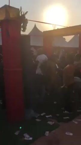 The video shows an outdoor party or celebration. People are gathered under a tent-like structure, with scattered money on the ground. A musician plays the drums and another person operates a DJ setup with turntables and a mixer. There are tables with drinks and food visible. The atmosphere appears lively and festive.