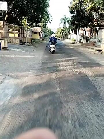 A person rides a white scooter down a paved road lined with trees and buildings. The road appears to be in a rural or suburban area. The sky is blue and clear, suggesting it is daytime. There is another person on a scooter further down the road. The camera is positioned behind the first scooter, giving a first-person perspective.