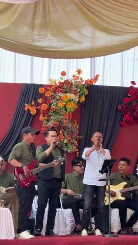 Here's a description of the video:

A band is performing live music on a stage in what appears to be an outdoor wedding reception area. There are six band members visible: two singers (one lead, one backing vocals), a bassist playing a red bass guitar, a drummer playing a compact set, and two guitarists playing electric guitars. All band members are dressed in green shirts, except for the lead singer, who is wearing a striped white shirt and black jeans. The backing vocalist is also wearing a black vest.

In front of the band, a woman wearing a hijab sits, likely a guest at the reception. The stage backdrop is a combination of red and black draping, adorned with a large arrangement of orange and red flowers. The stage is covered by a tent or canopy-like structure made of light-colored fabric.

The overall setting suggests a festive and celebratory atmosphere.