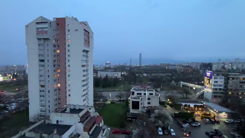 The video presents a high-angle cityscape taken during early evening or dusk. Dominating the foreground is a tall, rectangular residential building, light gray with pinkish accents, marked with the number 16. Some windows show interior lights on. Beneath it are lower structures with flat roofs. A green patch of grass and bare trees are visible between buildings. In the distance, a skyline shows several buildings, including a prominent, very tall skyscraper. The sky is overcast, and the light is muted. The video appears to be shot from a stationary position.