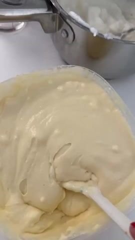 This media file features a close-up shot of a pale yellow, creamy substance being stirred with a white spatula in a clear plastic container. The substance appears thick and smooth, with some small air bubbles visible. In the background, a stainless steel pot with a similar-looking white substance is partially visible. The lighting suggests an indoor setting, likely a kitchen, with the white background indicating a clean surface.

The activity shown is the stirring or mixing of the creamy substance. There are no people clearly visible in the frame, though a hand holding the spatula is implied. The focus is entirely on the food preparation or creation process. The lack of external environmental cues makes it difficult to determine the time of day or weather. No text is visible in the media file. The context of Riihimäki, Finland, does not provide any specific visual cues within the frame itself.