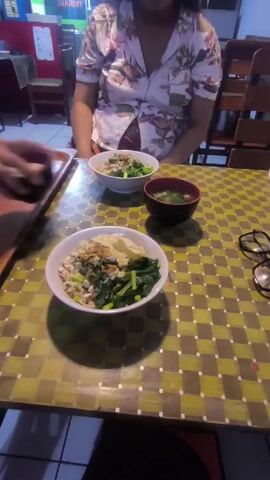 A person in a white floral shirt is sitting at a table with three bowls of food. The food appears to be a noodle dish with green vegetables and a light-colored broth. One bowl is being moved by a hand that is not the person's. There is a pair of glasses on the table, as well as a brown wooden tray. The table has a yellow and green checkered tablecloth. The background shows a restaurant setting with chairs and a sign.