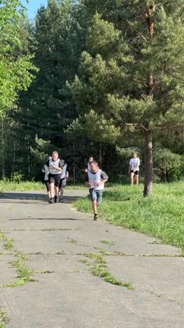 In a sunlit outdoor setting in Oreshniki, Russia, a group of young people, likely boys, are engaged in a running event. They are seen running on a worn paved path, which has some cracks and weeds growing through it. Lush green grass and numerous trees, including prominent pine trees, line the path, suggesting a park or forest environment.

The main subjects are several boys wearing white bibs with numbers. The boy in the foreground is wearing a bib that clearly displays the word "SLAVIC" and a number, possibly "5" or "8" (it's partially obscured). Another boy further back has a visible number "3" on his bib. They appear to be running with effort, indicating a race or a timed run. In the background, other individuals, including adults, can be seen standing or walking, possibly supervising or spectating. The weather appears clear and sunny, casting distinct shadows.
