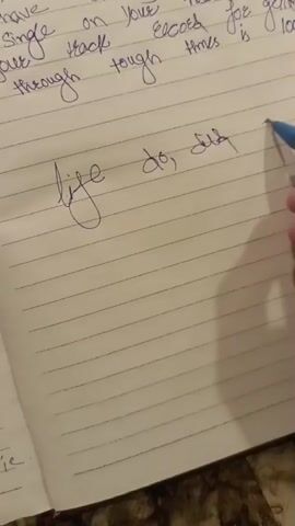 The video shows a person's hand writing in a notebook. The writing is in cursive and in blue ink. The notebook is lined and appears to be a standard composition notebook. The person is writing in the middle of the page. The top of the page has text that appears to be a motivational quote. The person's hand is holding a blue pen and is in the process of drawing a scribble on the right side of the page. The lighting is dim, suggesting the video was taken indoors with artificial light.