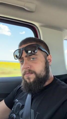 This is a close-up, selfie-style video taken from inside a vehicle. The subject, a man with dark hair, a beard, and sunglasses perched on his head, is looking directly at the camera. He is wearing a black t-shirt and a grey seatbelt is visible across his chest. Through the car window to his left, a blurry, yellow and green landscape is visible, suggesting the vehicle is in motion. The sky above is blue with scattered white clouds.