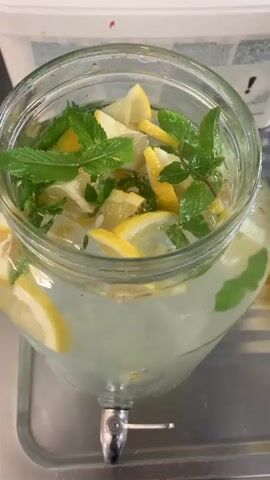 A close-up shot shows a glass beverage dispenser filled with lemonade, ice, lemon slices, and mint leaves. A person's hand, with neatly manicured nails, is seen stirring the contents with a metal spoon. The spoon is partially submerged in the drink, gently mixing the ingredients. The dispenser is placed on a metal tray, and in the background, a white container with bread slices is visible. The lighting is bright, illuminating the refreshing drink.
