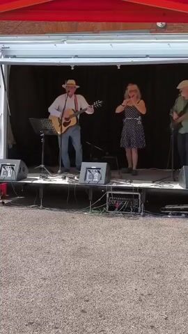 A band performs on an outdoor stage under a red awning. The stage is covered with a black backdrop. Three musicians are visible. On the left, a man in a white shirt, blue jeans, suspenders, and a straw hat plays an acoustic guitar and sings. In the center, a woman in a black and white polka dot dress plays a flute and sings. On the right, a man in a green shirt and a cap plays a stringed instrument, possibly a bass guitar. Two speakers and audio equipment are placed on the stage in front of the musicians. The ground in front of the stage is covered with gravel.