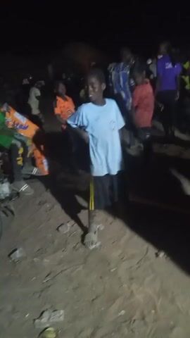 The video shows a group of people, mostly children, gathered in a dimly lit outdoor space. The ground appears to be dirt or sand. A boy wearing a light blue t-shirt and dark shorts is prominently visible in the foreground, dancing and moving around. Other children, wearing various colored shirts, are also present in the background. Some adults can also be seen in the crowd. The lighting is poor, and the scene appears to be captured at night. There is music playing, suggesting some form of gathering or celebration.