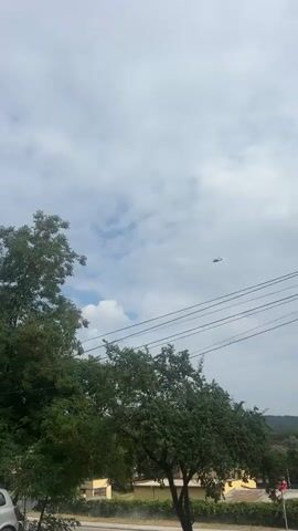 This video shows a helicopter flying in a cloudy sky. The helicopter is small and dark, appearing as a silhouette against the lighter sky. There are trees in the foreground, on the left and bottom of the frame, and power lines running horizontally across the lower part of the image. The helicopter is positioned in the upper right quadrant of the frame and moves slightly to the left and down as the video progresses. The sky is mostly blue with scattered white clouds.
