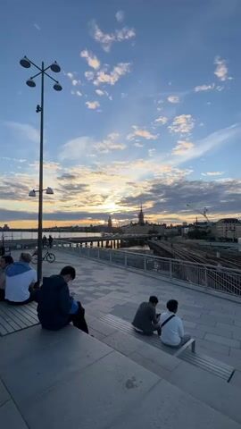 The video captures a serene sunset over Stockholm, Sweden. The sky is a canvas of blues and oranges, dotted with dramatic clouds that are catching the last rays of the sun. The iconic Stockholm City Hall, with its distinctive tower, is silhouetted against the vibrant sky, a prominent landmark in the city's skyline. A bridge crosses the water in the foreground, with traffic visible on it, suggesting the city is alive and bustling even as the day winds down.

In the foreground, several people are seated on tiered steps, enjoying the view. Some are facing the sunset, while others are engaged with their phones or conversing quietly. Their relaxed postures and the overall ambiance of the scene convey a sense of peace and appreciation for the beautiful twilight. The setting is an elevated walkway or plaza, offering an unobstructed panorama of the water and the cityscape. A modern lamppost stands tall on the left, adding a contemporary element to the scene.

The time of day is clearly evening, as indicated by the sunset and the lighting. The weather appears pleasant, with the clouds adding depth and visual interest to the sky without obscuring the sun entirely. The reflections of the city lights and the sky on the water create a shimmering effect, enhancing the picturesque quality of the moment. The scene evokes a feeling of calm, contemplation, and the simple joy of witnessing a beautiful natural spectacle in an urban setting. There is no visible text in the video.