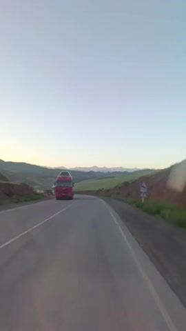 A truck is driving on a road. The truck is red and has a trailer. The truck is driving on a road with two lanes. The road is paved with asphalt. There are hills and mountains in the background. The sky is clear and blue. There are some trees and bushes on the side of the road. The video is shot from the perspective of another vehicle.