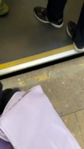 The video shows a low-angle view of a train platform.  Footwear of multiple people is visible, including sneakers and a lime green shoe. A light pink purse sits on the platform floor. A yellow safety line marks the edge of the platform. The flooring is a mix of textures, including concrete tiles and a textured gray surface. The scene suggests people are waiting on a train platform.