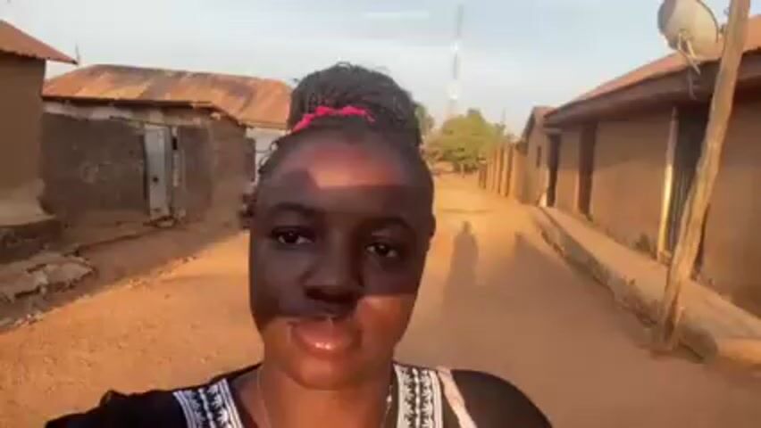 A young woman with dark skin is walking down a dirt road between modest buildings, recording a video. She has her hair in a braided bun with a red scrunchie. She is wearing a black top with white detailing around the neckline. The buildings on either side of the road appear to be constructed of mud or clay bricks with corrugated metal roofs. The sky is light blue. There is a telephone pole and a satellite dish visible in the background.