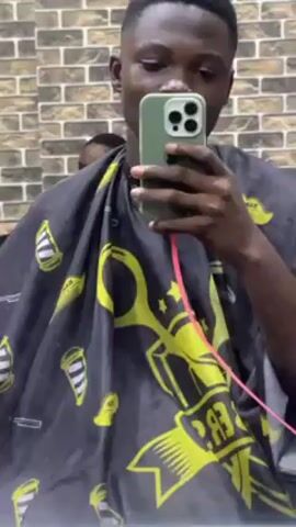 A young man stands in front of a mirror, holding an iPhone 13 or 14, taking a selfie. He is wearing a black barber's cape with yellow designs. He has short, dark hair. Behind him is a brick wall and another person getting a haircut. The audio suggests he is at a barber shop and inquiring about the cost of a hair service.