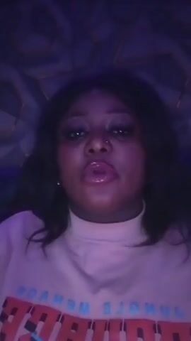 A woman is indoors, likely in a club. She is wearing a light-colored turtleneck shirt and has long, dark hair. The lighting is dark with a purple hue. There is a patterned wall behind her. She touches her face and makes faces, possibly in time with the music. Loud music is audible in the background.