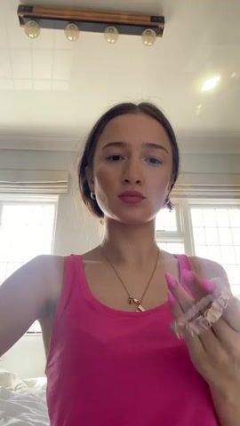 A young woman with long dark hair and bright pink fingernails, wearing a bright pink tank top and a gold necklace with two charms, is holding two bracelets in her hands. She is looking directly at the camera. The video appears to be filmed indoors, with a ceiling light fixture visible overhead and a window with blinds in the background. The woman is in the process of putting on a bracelet.