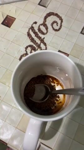 A close-up overhead shot shows a white mug on a checkered surface. Inside the mug, a spoon rests on a small amount of dark liquid and instant coffee granules. Hot water is poured into the mug, dissolving the coffee and creating a foamy layer as it fills the cup. The surface beneath the mug has a decorative pattern of coffee beans arranged to form the word "coffee."