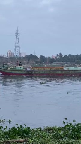 The video shows a green and red barge moving across a body of water. There are people visible on the barge. The background includes a power line tower, buildings, trees, and a cloudy sky. Water hyacinths are visible near the bank of the waterway. The barge appears to be moving from left to right in the frame.